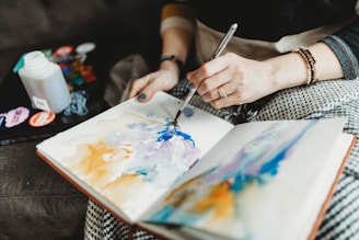 A person is painting in a notebook using watercolors. The artwork features abstract swirls of blue, purple, and orange. The individual holds a brush in one hand and is wearing a ring and bracelets on the other. There are art supplies and a bottle behind the hands, suggesting a creative workspace.