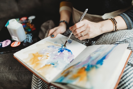 A person is painting in a notebook using watercolors. The artwork features abstract swirls of blue, purple, and orange. The individual holds a brush in one hand and is wearing a ring and bracelets on the other. There are art supplies and a bottle behind the hands, suggesting a creative workspace.