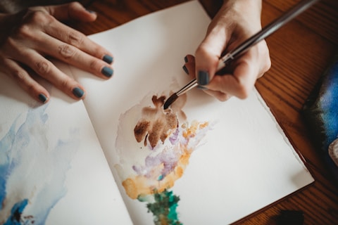 A person is painting with watercolors on a sketchbook, using a brush to create an abstract design with shades of brown, purple, and green. Their nails are painted a dark blue, and the scene is set on a wooden table with a fabric pouch visible in the corner.