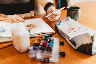 A person sits at a wooden table engaged in a creative activity involving watercolors. Open sketchbook with painted artwork, a hand holding a pen, a small watercolor palette, a cup, and a pouch with floral design are visible. The scene suggests a peaceful, artistic environment.
