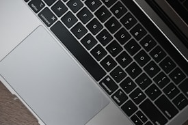 A close-up view of a laptop keyboard and touchpad, featuring neatly arranged black keys with white lettering on a gray metallic surface, set against a wooden background.