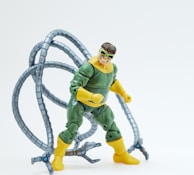 An action figure resembling a superhero or villain character with mechanical tentacles attached to its back. The figure is dressed in a green and yellow suit, wearing green goggles, and positioned in a dynamic pose.