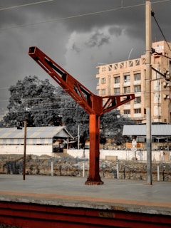 A railway station platform with a large red metal structure in the center. There is an old, beige building with a sign that reads 'HOTEL' in the background. Trees and power lines are visible, and the sky appears dark and cloudy, possibly indicating an impending storm. The platform is mostly empty with a few small structures, including a shed-like building.