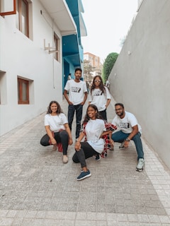 A group of five individuals are positioned in an outdoor alley, framed by modern buildings with a blue accent wall. They are casually posed, wearing matching white t-shirts with black text, displaying a relaxed and friendly demeanor.