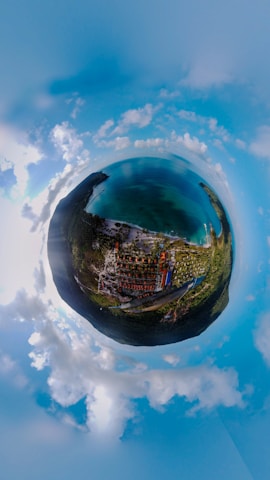 A panoramic aerial view showcasing a lush green landscape and a turquoise body of water, creating a tiny planet effect. Below, red-roofed structures and a network of roads are visible amidst tropical vegetation. White clouds are scattered across a bright blue sky.