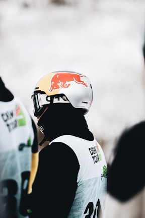 A person wearing a helmet with a prominent Red Bull logo, dressed in ski gear including a white jacket with 'Dew Tour' written on it. The individual appears to be participating in a winter sports event.