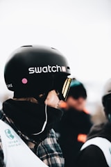 A person is standing or sitting, wearing a black helmet branded with the Swatch logo. They have a plaid jacket and a scarf or face covering around their neck. Visible on their outfit is a Copper Mountain logo. In the blurred background, other individuals can be seen, likely in a snowy or cold environment.