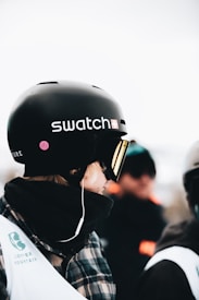A person is standing or sitting, wearing a black helmet branded with the Swatch logo. They have a plaid jacket and a scarf or face covering around their neck. Visible on their outfit is a Copper Mountain logo. In the blurred background, other individuals can be seen, likely in a snowy or cold environment.