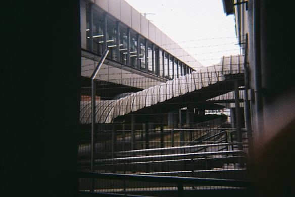 A view of an urban infrastructure featuring a pedestrian bridge covered with protective awnings. Barbed wire and metal fences are in the foreground, suggesting a secure or restricted area. The background shows a modern building with large glass windows.