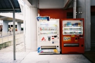 Two vending machines are placed side by side under a sheltered area. The machine on the left is white, stocked with beverages, and displays various drinks through a glass front. The right machine is orange with visible branding, also filled with bottles and cans. Some people are walking in the background near a fenced pathway.