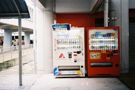 Two vending machines are placed side by side under a sheltered area. The machine on the left is white, stocked with beverages, and displays various drinks through a glass front. The right machine is orange with visible branding, also filled with bottles and cans. Some people are walking in the background near a fenced pathway.