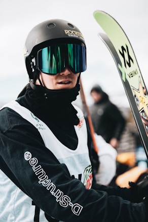A person wearing a helmet and reflective ski goggles holds a pair of skis. The individual is dressed in a white vest and black clothing underneath. The helmet features a logo, and there are various logos on the clothing and equipment. The background is slightly blurred, suggesting a snowy or wintry setting.