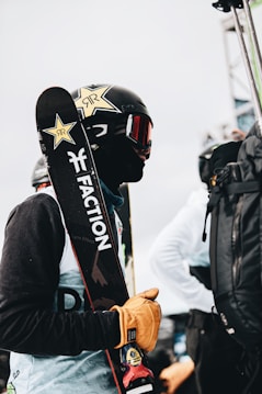 A person is dressed in winter sports gear, holding a pair of skis over their shoulder. The individual is wearing a black helmet with a star design and ski goggles, and is also outfitted with gloves. The environment appears to be an outdoor setting, possibly at a ski resort or event.