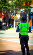 A security officer wearing a fluorescent yellow vest is standing on a paved walkway. The background shows a street with colorful storefronts and green trees, implying an urban or town setting.