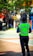 A security officer wearing a fluorescent yellow vest is standing on a paved walkway. The background shows a street with colorful storefronts and green trees, implying an urban or town setting.