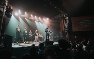 A band performs on stage in front of an audience, with bright stage lights overhead and a screen displaying the song lyrics. Several musicians play instruments like guitar and keyboards, while a lead singer engages with the crowd.