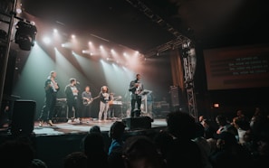 A band performs on stage in front of an audience, with bright stage lights overhead and a screen displaying the song lyrics. Several musicians play instruments like guitar and keyboards, while a lead singer engages with the crowd.