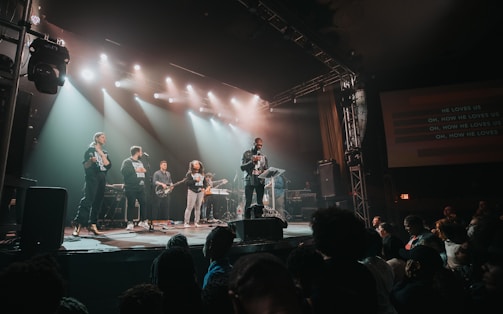 A band performs on stage in front of an audience, with bright stage lights overhead and a screen displaying the song lyrics. Several musicians play instruments like guitar and keyboards, while a lead singer engages with the crowd.