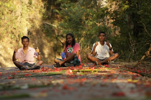 Three young people are sitting cross-legged on a path covered with scattered red petals and foliage. They are in a serene outdoor setting surrounded by greenery. The two individuals on the sides are giving thumbs-up gestures, while the one in the middle is smiling. The sunlight is casting warm tones across the scene.
