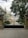 An ornate black wrought iron gate stands closed, leading to a grand building with classical architecture. The building features decorative details, with columns and light-colored stonework. Lush green trees and manicured bushes line the pathway leading to the building.