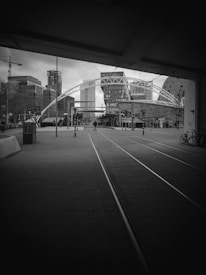 An urban scene featuring a modern architectural structure with a large archway over a pedestrian area. Skyscrapers and office buildings define the skyline in the background, while tram tracks run through the foreground. There are a few people walking in the distance and bicycles parked to the side.