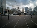 A modern urban scene with tram tracks leading through a street lined with contemporary buildings. In the background, a construction crane is visible against a cloudy sky, with several high-rise structures, including a distinctive curved building, marking the cityscape. The area appears open and spacious, with a mix of pedestrians and bicycles visible along the sides.