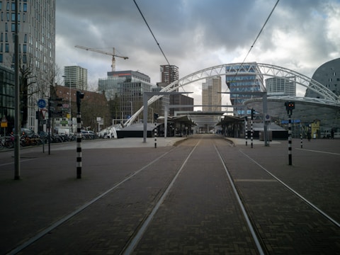 A modern urban scene with tram tracks leading through a street lined with contemporary buildings. In the background, a construction crane is visible against a cloudy sky, with several high-rise structures, including a distinctive curved building, marking the cityscape. The area appears open and spacious, with a mix of pedestrians and bicycles visible along the sides.