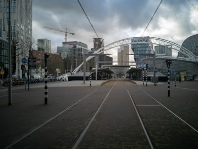 A modern urban scene with tram tracks leading through a street lined with contemporary buildings. In the background, a construction crane is visible against a cloudy sky, with several high-rise structures, including a distinctive curved building, marking the cityscape. The area appears open and spacious, with a mix of pedestrians and bicycles visible along the sides.