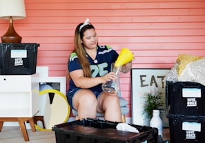 A person is sitting in a room with a pink wooden wall. They are wearing a sports jersey and headband, assembling a glass vase with a yellow funnel. The room contains various items including Hive Boxx storage bins, a lamp, pillows, a framed art piece with the word 'EAT', and a small plant. A small table and a round mirror are also visible.