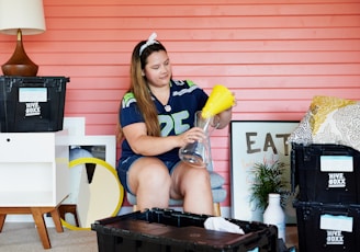 A person is sitting in a room with a pink wooden wall. They are wearing a sports jersey and headband, assembling a glass vase with a yellow funnel. The room contains various items including Hive Boxx storage bins, a lamp, pillows, a framed art piece with the word 'EAT', and a small plant. A small table and a round mirror are also visible.