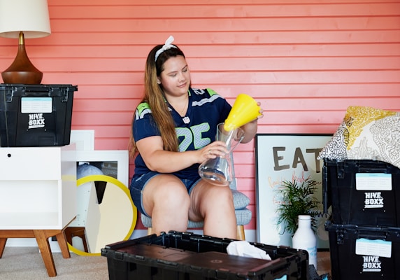 A person is sitting in a room with a pink wooden wall. They are wearing a sports jersey and headband, assembling a glass vase with a yellow funnel. The room contains various items including Hive Boxx storage bins, a lamp, pillows, a framed art piece with the word 'EAT', and a small plant. A small table and a round mirror are also visible.
