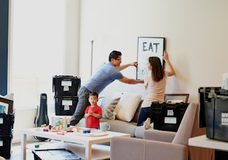 A couple is setting up a new home by hanging a decor piece on the wall while a child stands nearby. Various moving boxes and a guitar case are scattered around the room. A low table is covered with children's toys, and a couch with cushions is positioned against the wall.