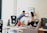 A couple is setting up a new home by hanging a decor piece on the wall while a child stands nearby. Various moving boxes and a guitar case are scattered around the room. A low table is covered with children's toys, and a couch with cushions is positioned against the wall.