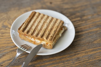 A grilled sandwich with defined grill marks is placed on a white plate. The sandwich appears to have a filling of melted cheese and possibly other ingredients. Next to the plate, on a wooden surface, there is a knife and fork.