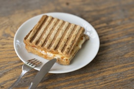A grilled sandwich with defined grill marks is placed on a white plate. The sandwich appears to have a filling of melted cheese and possibly other ingredients. Next to the plate, on a wooden surface, there is a knife and fork.