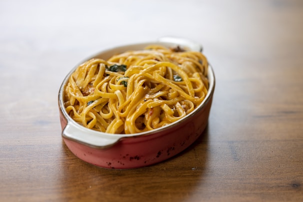 A ceramic dish filled with a serving of pasta, likely fettuccine, covered in a creamy sauce with visible herbs. The pasta is neatly arranged in the oval dish, which has handles on both ends.