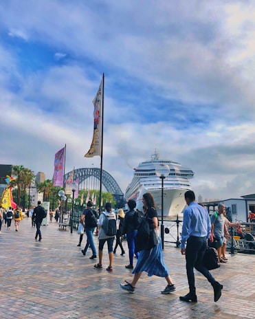 A bustling waterfront scene with people walking along a promenade. A large cruise ship is docked, and the iconic arch of a steel bridge is visible in the background. Various colorful flags line the walkway, creating a vibrant atmosphere. The sky is partially cloudy with hints of blue.