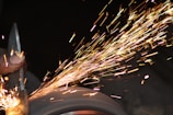 A hand is holding a piece of metal against a grinding wheel, producing a shower of bright, colorful sparks flying off into the dark background.