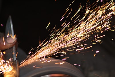 A hand is holding a piece of metal against a grinding wheel, producing a shower of bright, colorful sparks flying off into the dark background.
