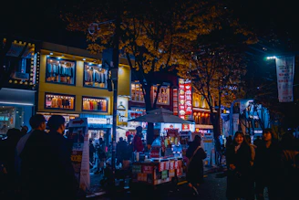 A nighttime cityscape with a lively food market bustling with people and twinkling lights.