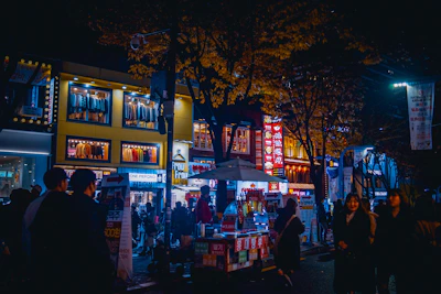 A nighttime cityscape with a lively food market bustling with people and twinkling lights.