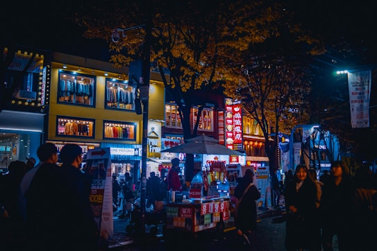 A bustling night scene features a lively street market with neon lights illuminating the area. Several people walk past various storefronts, with clothing displays visible through large windows above. Food carts and vendors line the street, creating a vibrant and dynamic atmosphere.