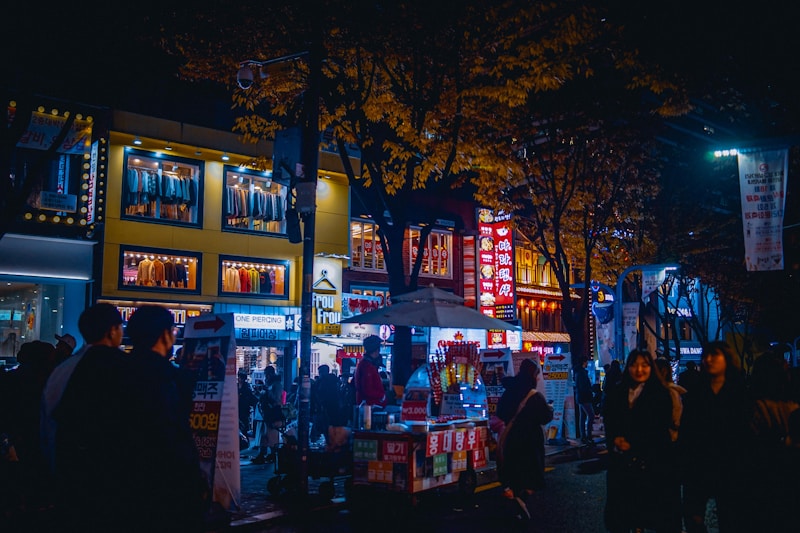 A bustling night scene features a lively street market with neon lights illuminating the area. Several people walk past various storefronts, with clothing displays visible through large windows above. Food carts and vendors line the street, creating a vibrant and dynamic atmosphere.