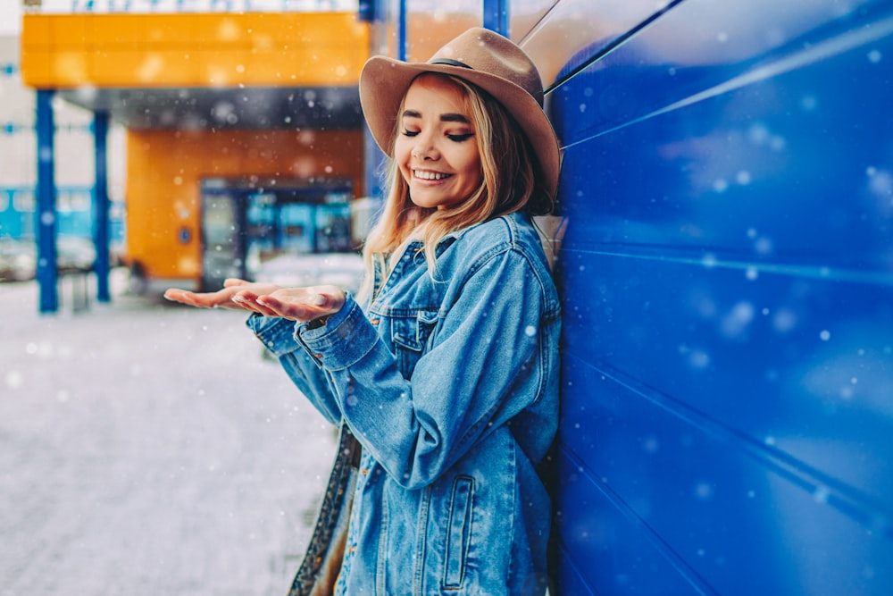 mulher na jaqueta jeans azul e chapéu fedora marrom em pé no chão coberto de neve durante o dia