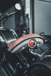 A close-up view of a motorcycle seat with a brown leather cover and a red brake light beneath it. The image captures part of the vehicle's handlebars and a mirror, and the background features a muted indoor setting.
