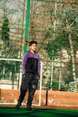 A young person stands on a sports field, wearing a purple and black tracksuit. The background features a wire fence and bare trees, suggesting an outdoor urban environment in a cooler season.