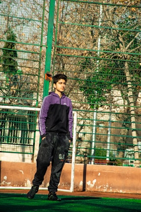 A young person stands on a sports field, wearing a purple and black tracksuit. The background features a wire fence and bare trees, suggesting an outdoor urban environment in a cooler season.