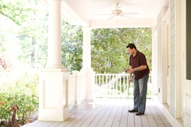 A man is using a hose to water or clean a porch area surrounded by greenery. The porch features white columns and railing, with a ceiling fan above. He is dressed casually in a dark polo shirt and jeans, and the environment is bright, suggesting a sunny day.
