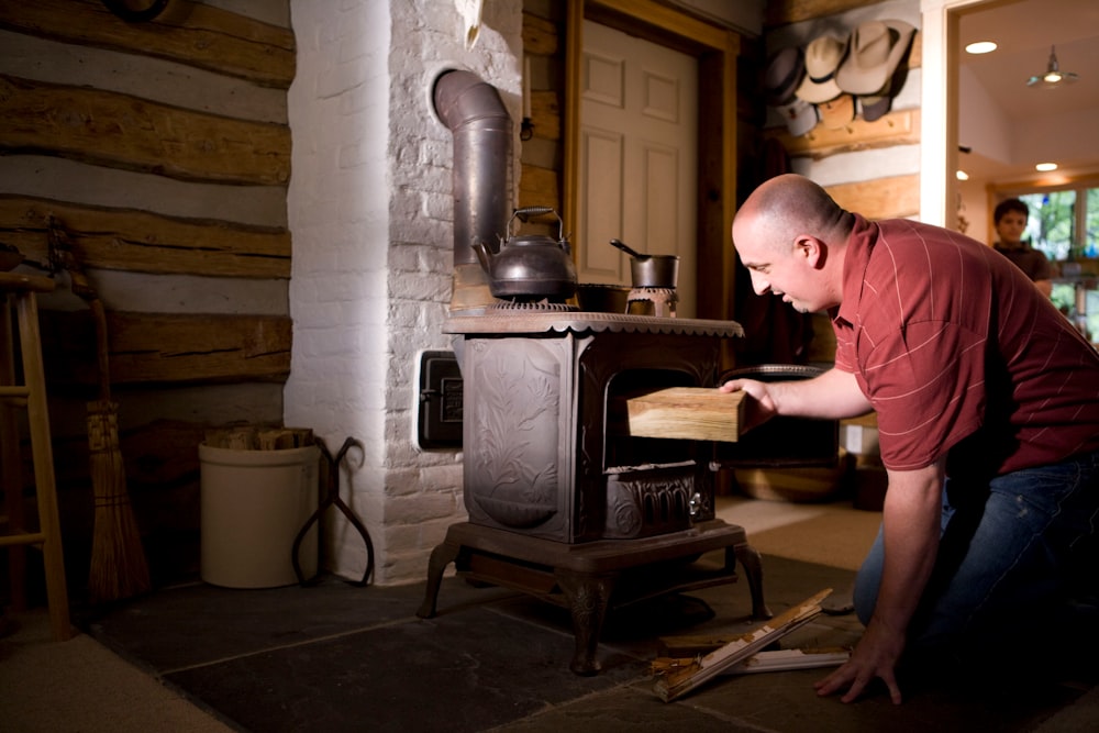 man in pink polo shirt and blue denim jeans standing in front of black wood burner