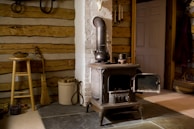 A rustic room featuring a traditional wood-burning stove made of cast iron, placed on a stone floor. The walls are constructed from wooden logs, giving a cabin-like feel. Nearby, a wooden stool supports a bowl, while a broom and a bucket filled with kindling rest against the wall. The room is warmly lit, with a door visible in the background.